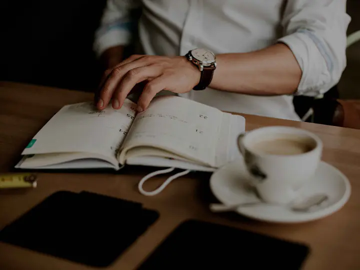 A hand resting on an open notebook beside a cup of coffee