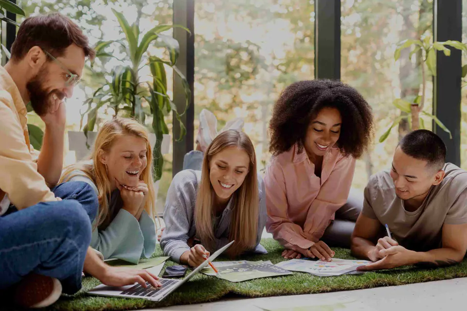 A group lying on a grass mat, laughing and working together on papers and a laptop