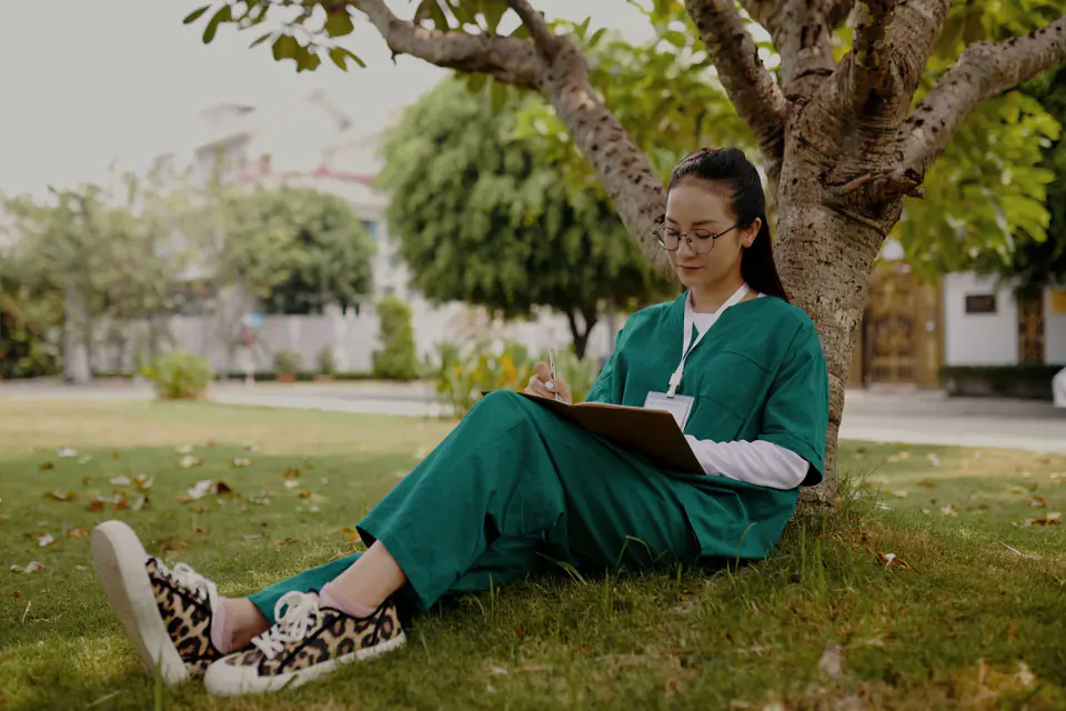 A therapist in green scrubs reading a book on a lawn under a tree