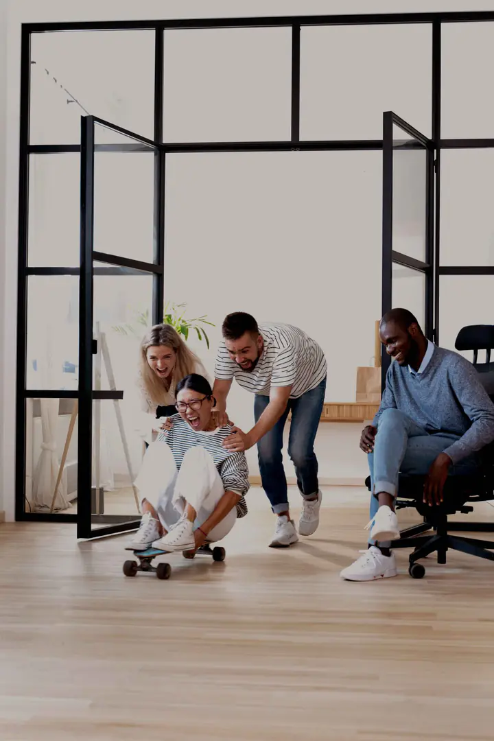 Colleagues laughing in an office as one is pushed along on a skateboard