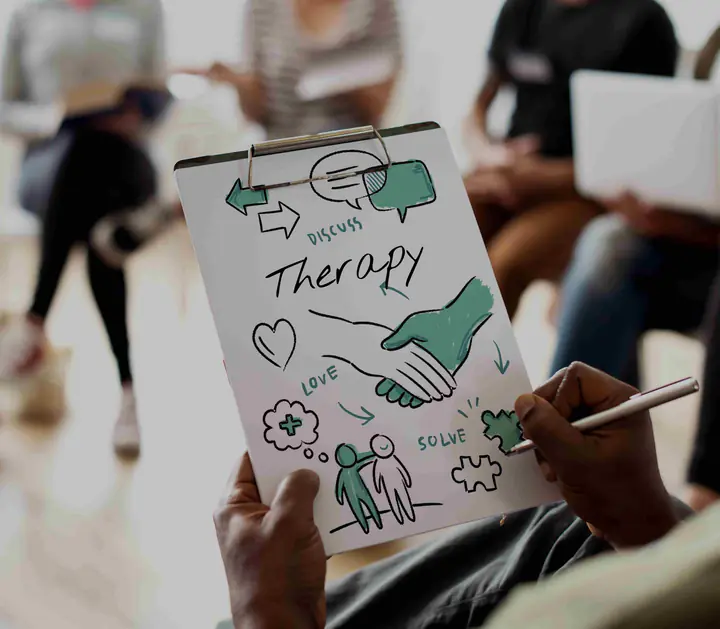 A hand drawing a sketch titled “Therapy” on a clipboard during a group session